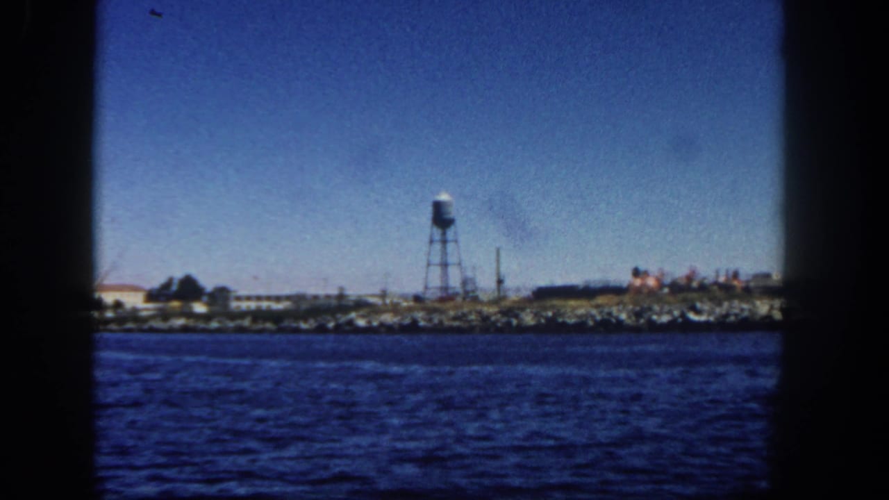San Diego waterfront in 1962, viewed from a moving vessel with a water tower, low industrial structures, and shoreline rocks.