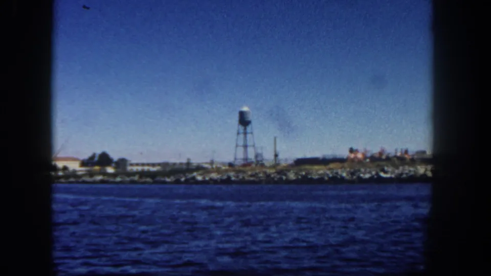 San Diego waterfront in 1962, viewed from a moving vessel with a water tower, low industrial structures, and shoreline rocks.