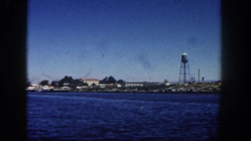 1962 vessel-side view of San Diego harbor area, with blue water, rocky shore, water tower, and possible cranes or masts.