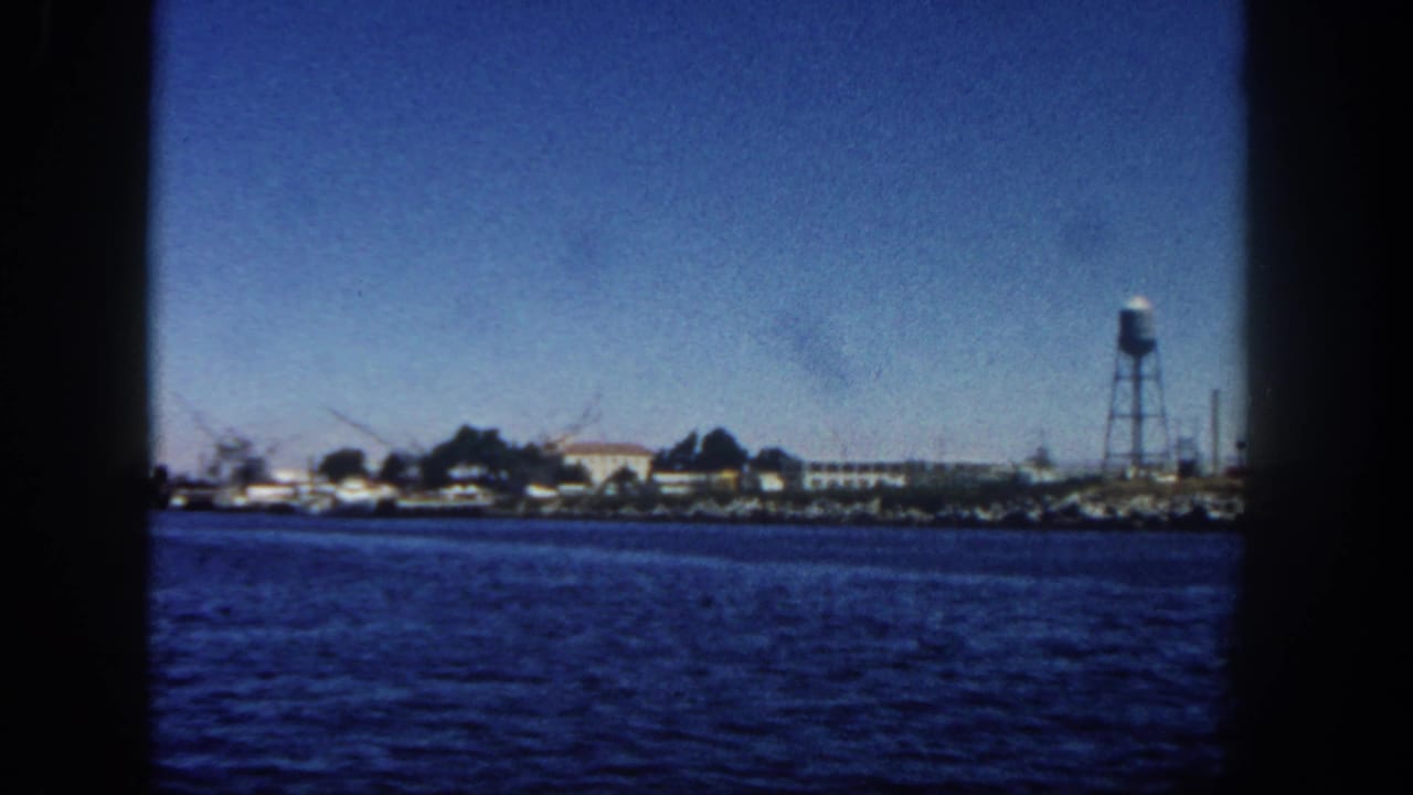 Blue harbor water with a distant shoreline, water tower, and low buildings under clear sky.