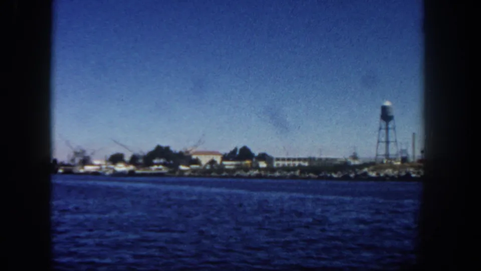 1962 San Diego waterfront seen from the water, with blue harbor foreground, distant buildings, cranes or masts, and a tall water tower.
