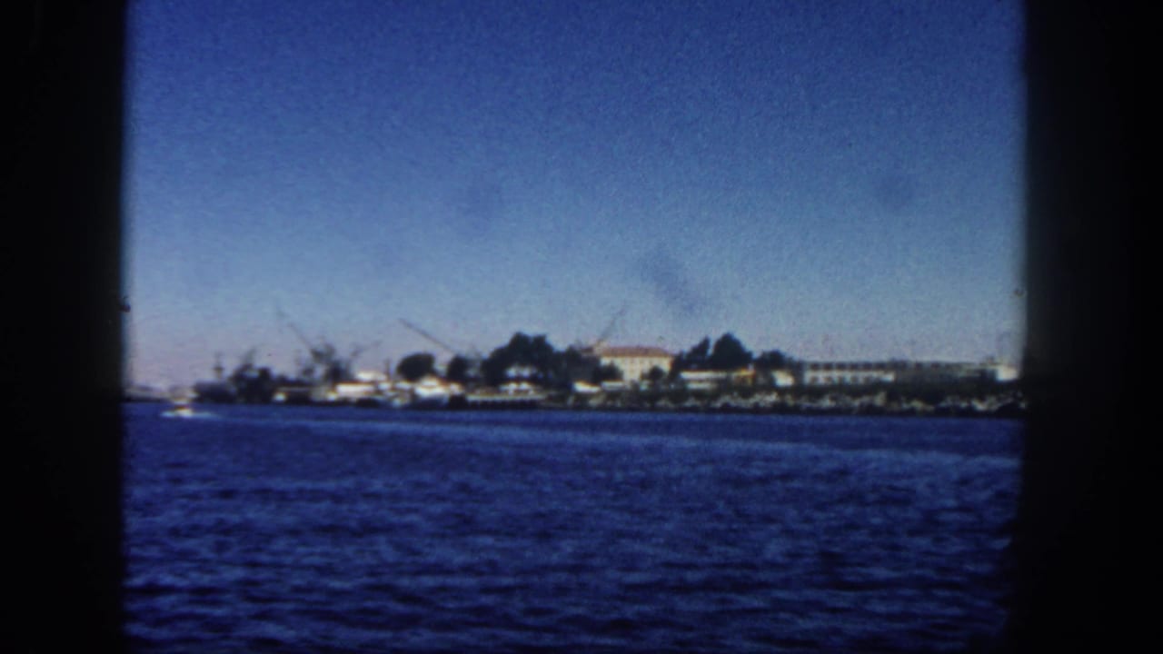 San Diego harbor view from a moving vessel, showing blue water, low shoreline structures, cranes or masts, and clear sky.