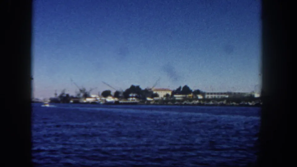 San Diego harbor view from a moving vessel, showing blue water, low shoreline structures, cranes or masts, and clear sky.