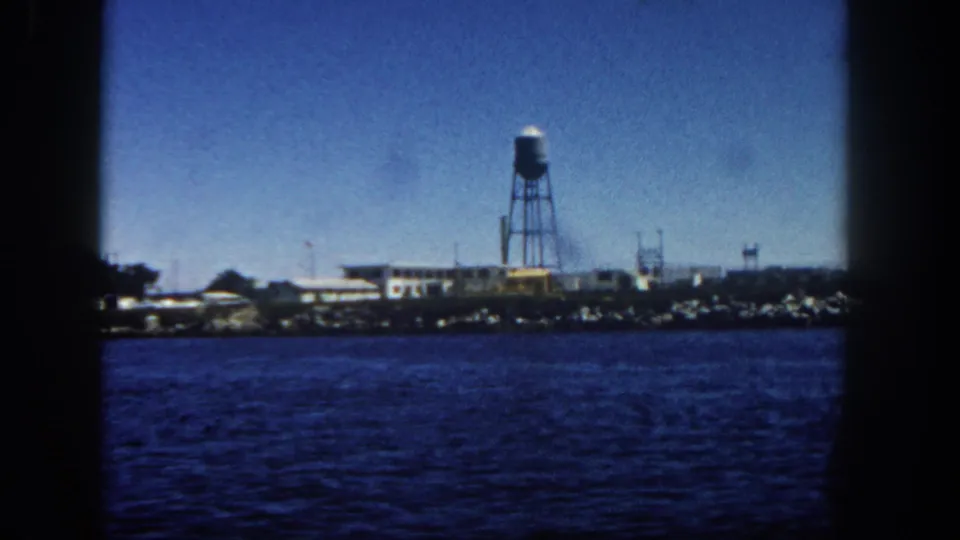 1962 San Diego shoreline with a prominent water tower and low waterfront buildings, viewed across rippled harbor water.