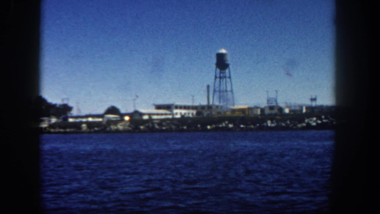 1962 San Diego waterfront seen from a ship, with rippling water, low buildings, a tall water tower, and dark frame edges.