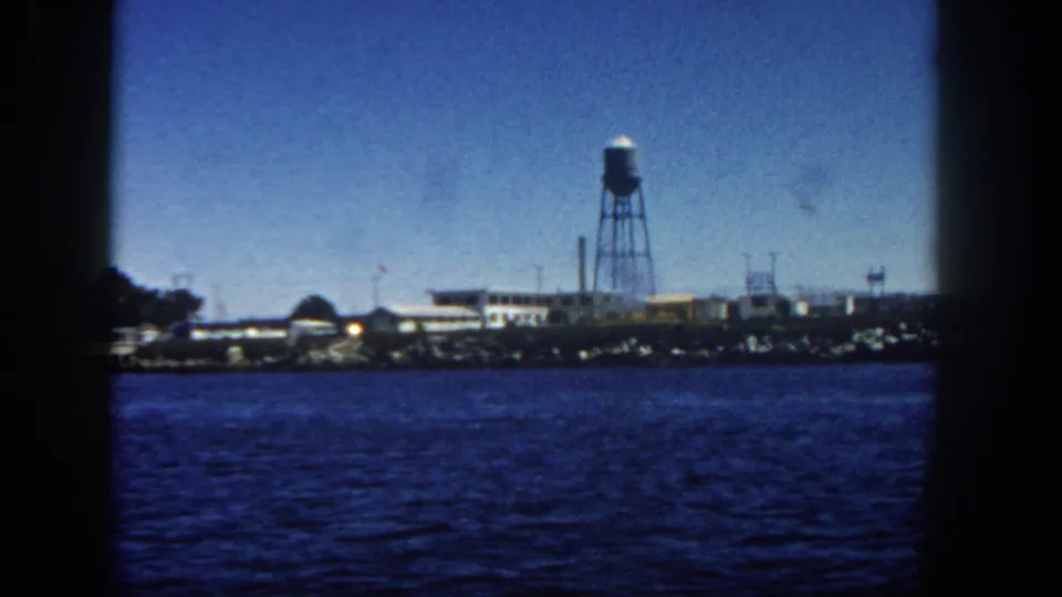 1962 San Diego waterfront seen from a ship, with rippling water, low buildings, a tall water tower, and dark frame edges.