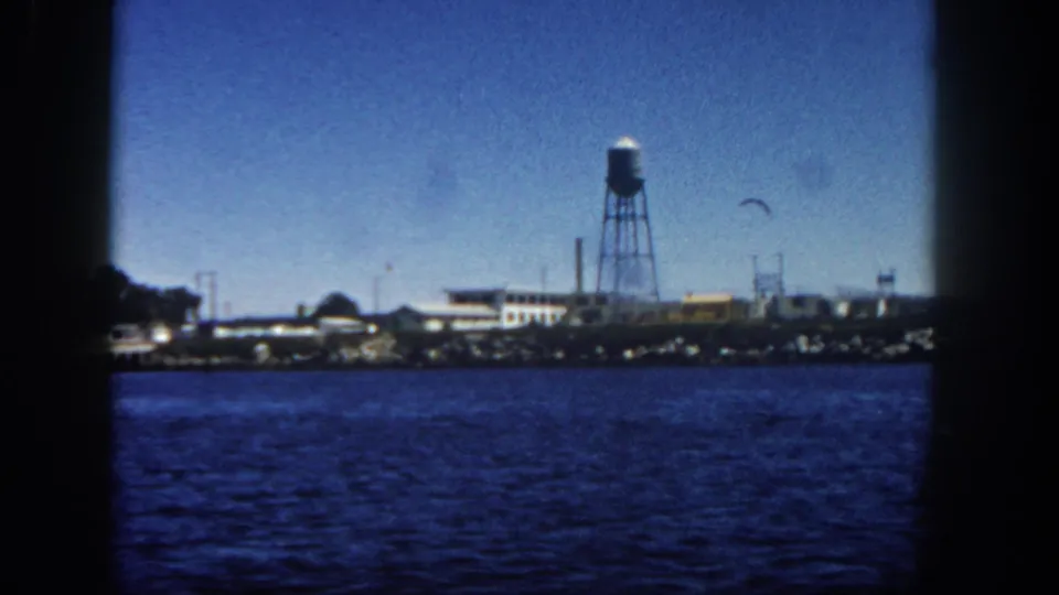 1962 San Diego harbor view with a distant bird, industrial buildings, rocky shoreline, and a tall water tower beyond blue water.