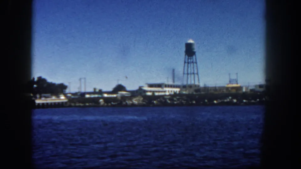 1962 San Diego industrial waterfront viewed across the harbor, with low buildings, a water tower, distant structures, and dark film-frame edges.