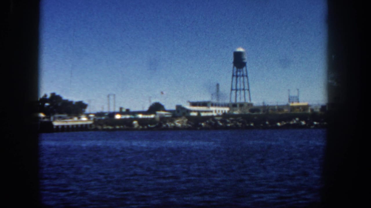 Harbor water with an industrial shoreline and a tall water tower in the distance.