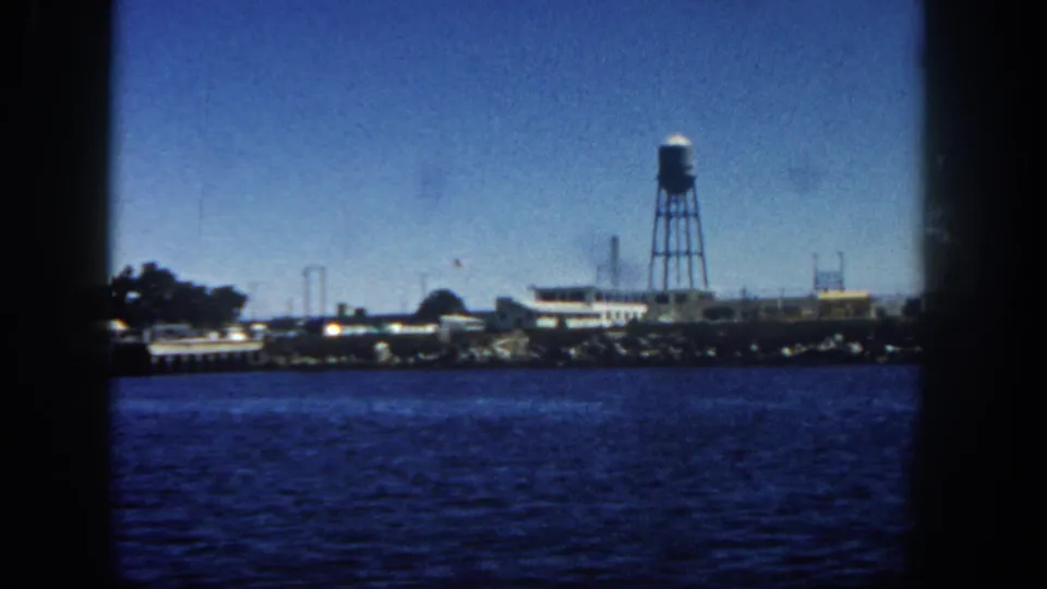 1962 San Diego harbor view from a boat, showing blue water, low waterfront buildings, and a tall water tower beyond the shoreline.