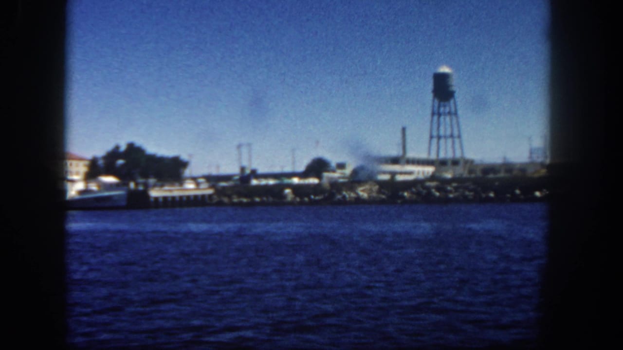 Industrial shoreline across blue water with a water tower near the right side of frame.