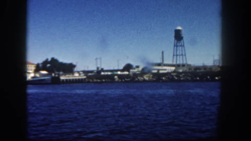 Boat-shot 1962 San Diego waterfront frame with a water tower, dockside buildings, and calm blue harbor water in front.