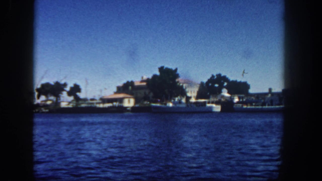 1962 San Diego bay view from an 8mm home movie, showing rippling water, distant waterfront buildings, trees, small boats, and harbor cranes.