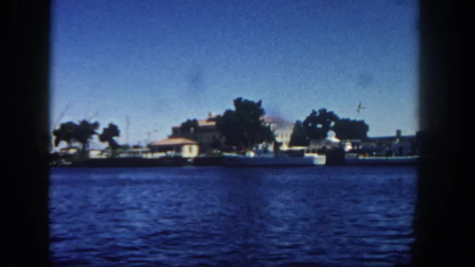 1962 San Diego bay view from an 8mm home movie, showing rippling water, distant waterfront buildings, trees, small boats, and harbor cranes.