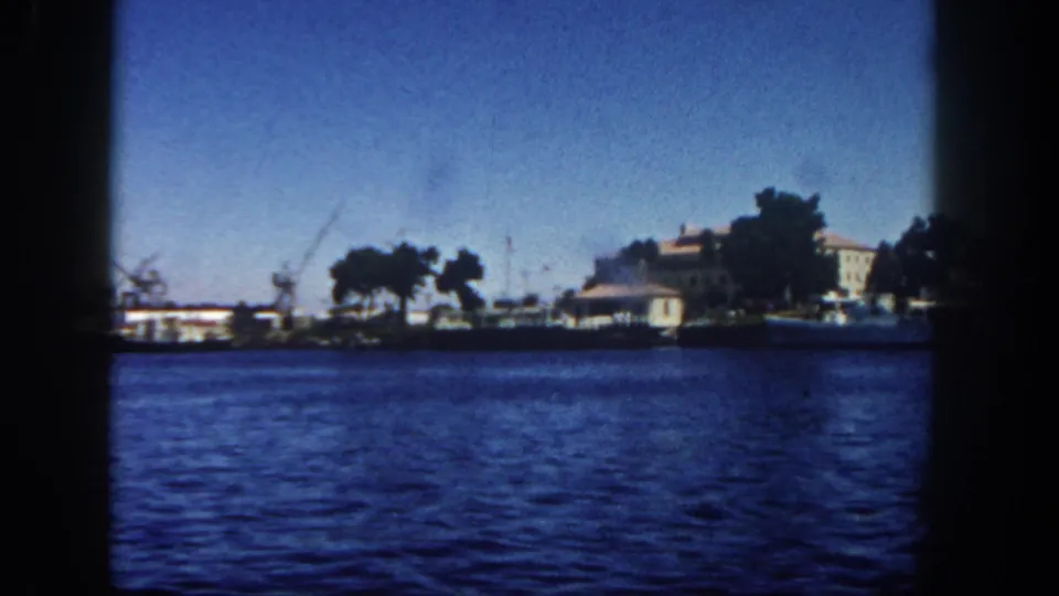 Moving-boat view of a San Diego waterfront in 1962, showing cranes, low buildings, trees, boats, and textured blue bay water.