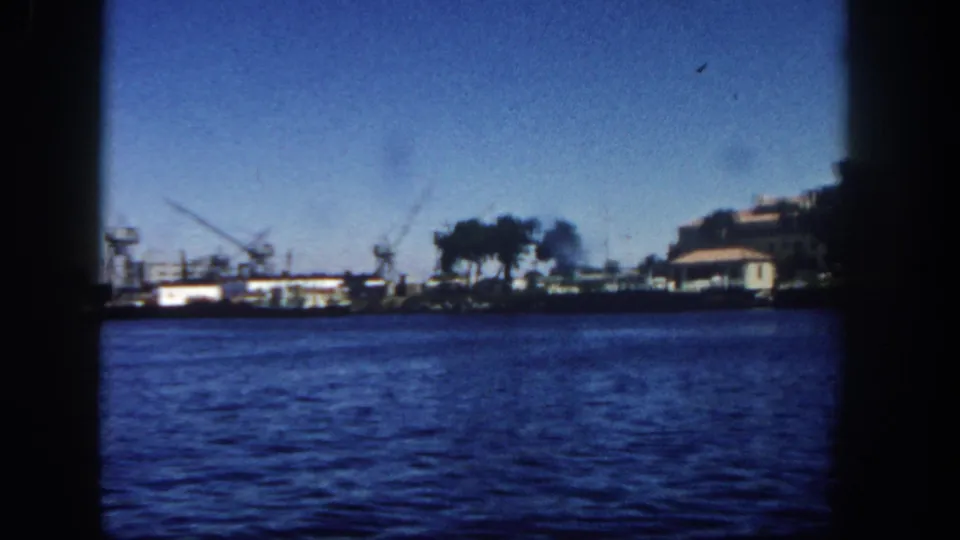1962 San Diego bay footage showing a wider shoreline view with cranes, trees, boats, low waterfront buildings, and saturated blue water in the foreground.
