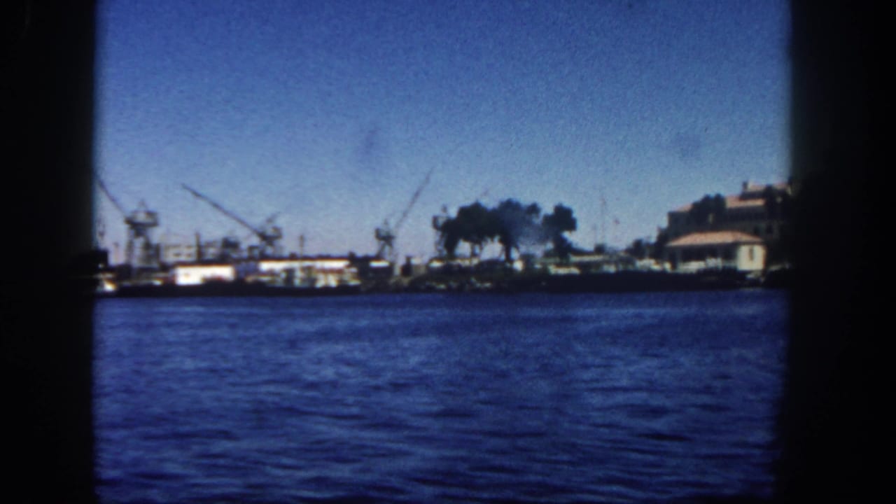 Harbor water with distant cranes, industrial buildings, smoke, and dark film-frame edges.