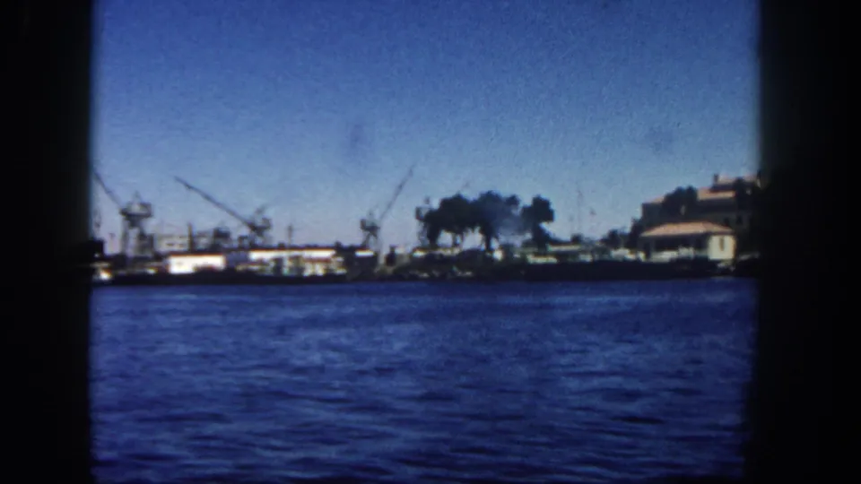 1962 San Diego harbor view from a boat, showing blue water, distant dock cranes, industrial shoreline, smoke, and dark 8mm frame edges.
