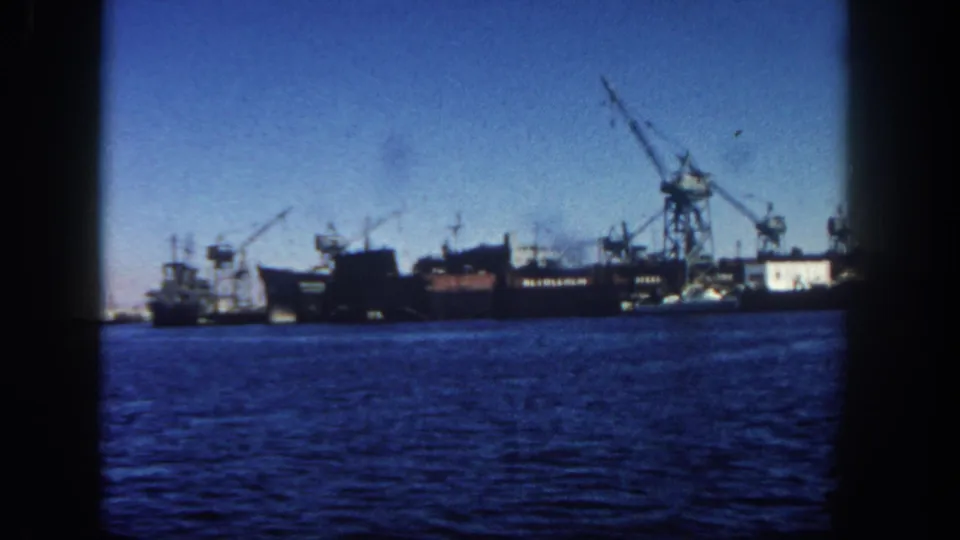 1962 San Diego harbor view from the water, with a ship, dock cranes, smoke, industrial buildings, and dark 8mm frame borders.