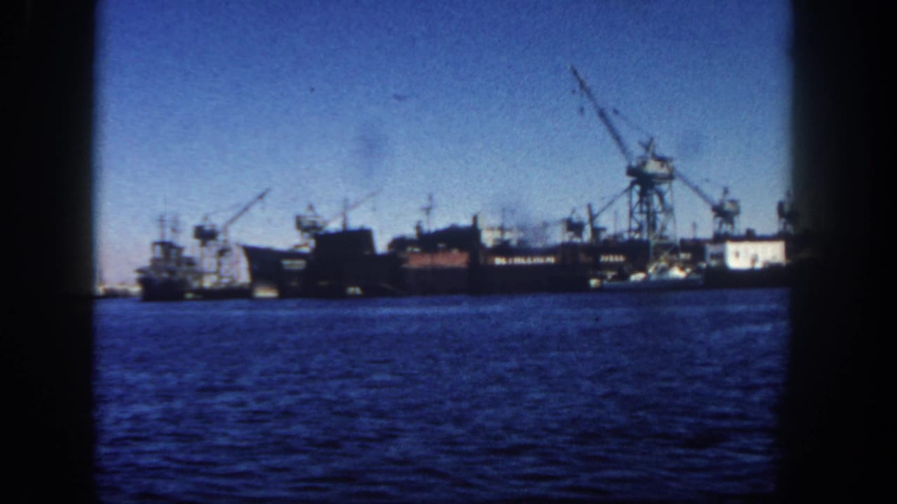 Distant ships and cranes across blue harbor water under a clear sky.