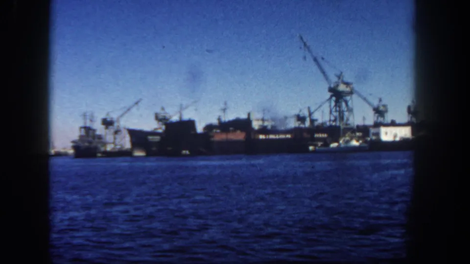 1962 San Diego harbor view with blue water in the foreground and docked ships and cranes along the distant waterfront.