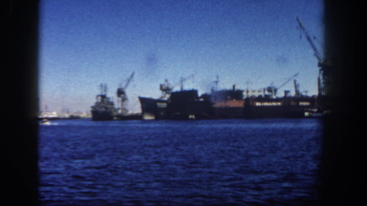 Large docked vessel and cranes across blue harbor water with dark film borders.