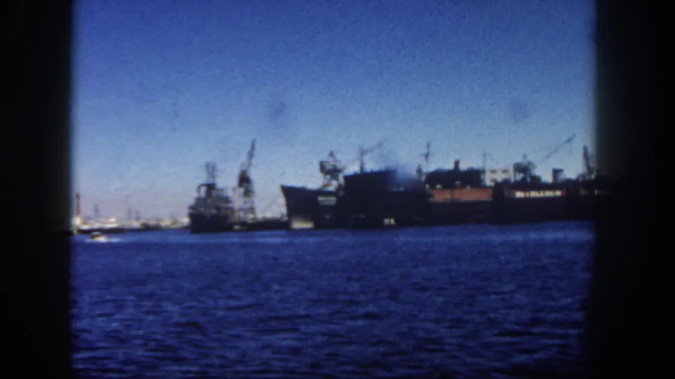 Across blue San Diego harbor water, a large docked vessel sits near cranes and low industrial waterfront buildings.