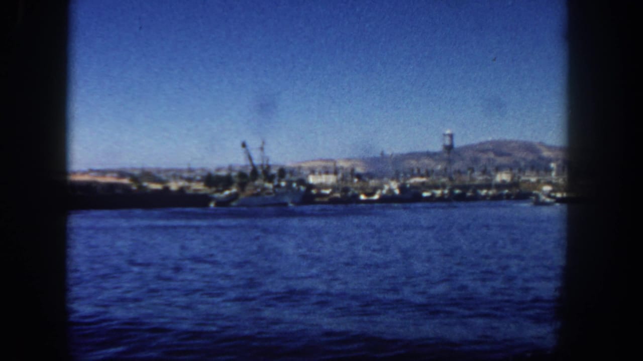 Harbor water facing a distant shipyard with cranes, vessels, hills, and a water tower.