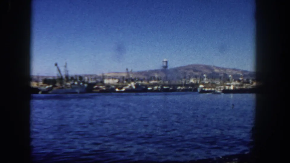 1962 home-movie view of San Diego's working harbor, with blue water in front and cranes, vessels, industrial structures, and hillside beyond.