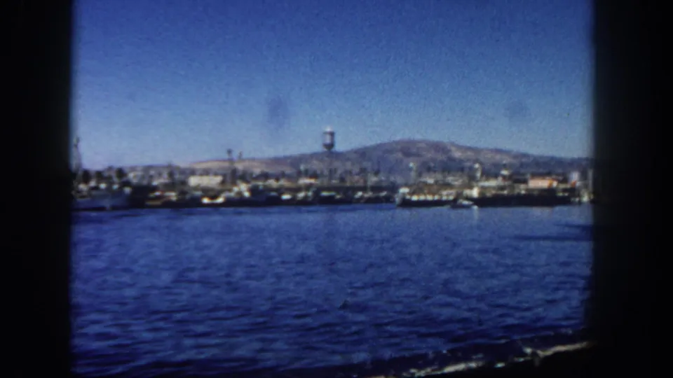 Wide 1962 San Diego harbor frame showing calm water, distant industrial docks, cranes, moored vessels, hillside terrain, and a water tower.