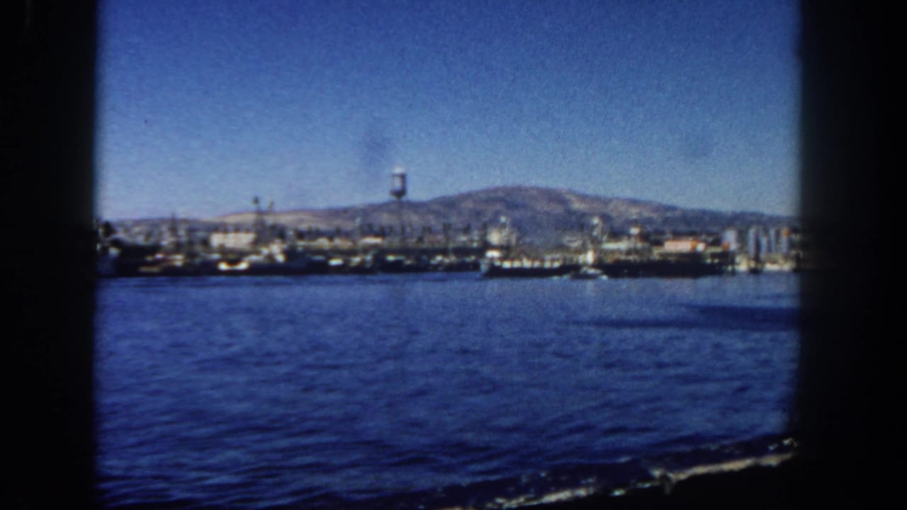 Harbor water with docked ships, a water tower, shoreline buildings, and mountains beyond.