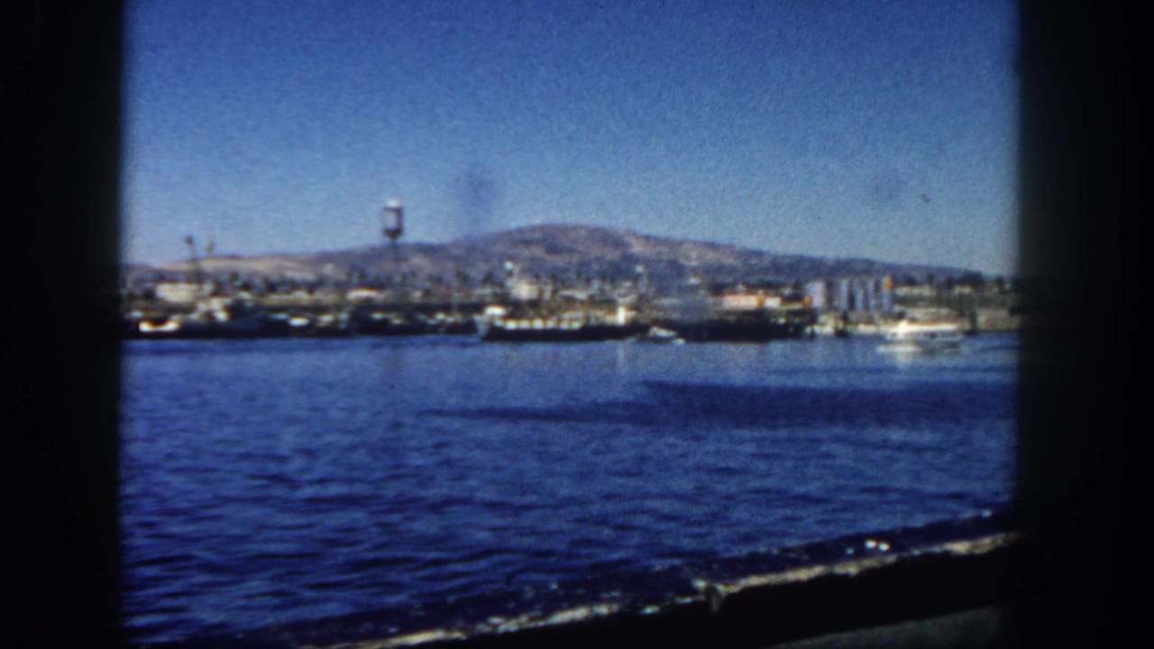 Wide harbor view with ships along the shore, a water tower, mountains, and a small boat at right.