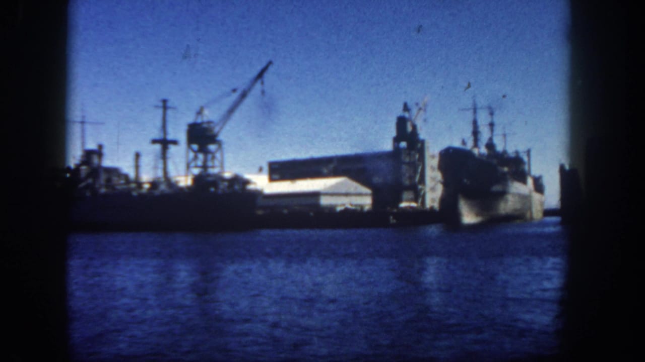 Naval ship docked beside cranes and waterfront buildings across blue harbor water.