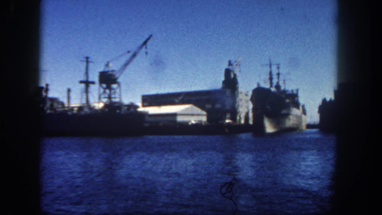 Harbor view of docked naval vessel, crane, warehouse roof, and water in foreground.