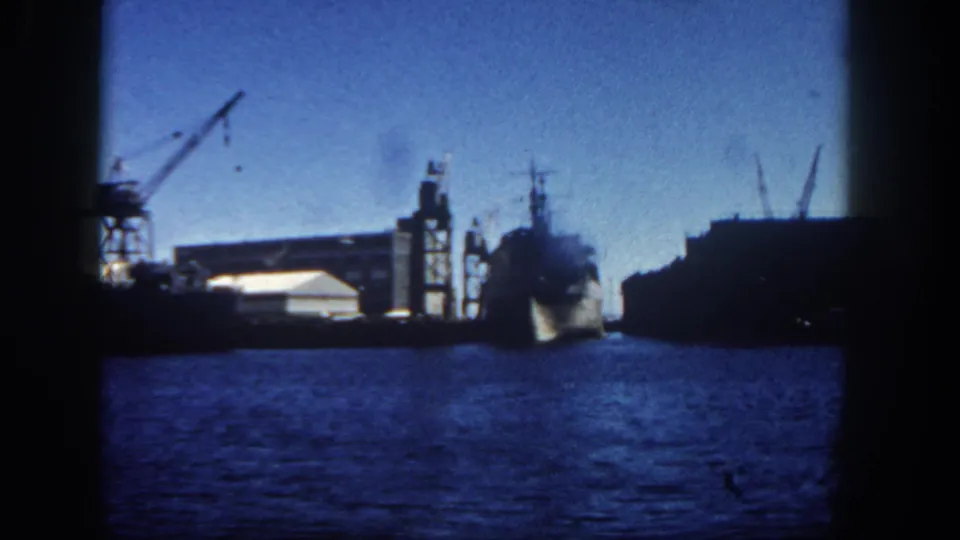 San Diego harbor footage showing a naval ship near silhouetted dock structures, cranes, and rippling water.