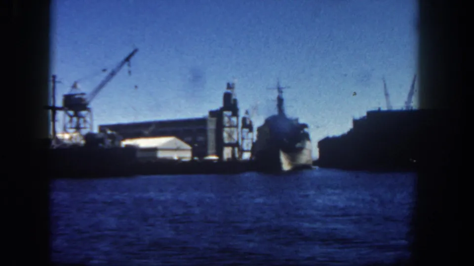 1962 San Diego harbor view across blue water toward docked industrial vessels, cranes, and waterfront structures on 8mm film.