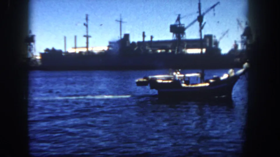 A small boat moves across San Diego harbor water in front of docked industrial ships, cranes, and waterfront structures in 1962 home-movie footage.
