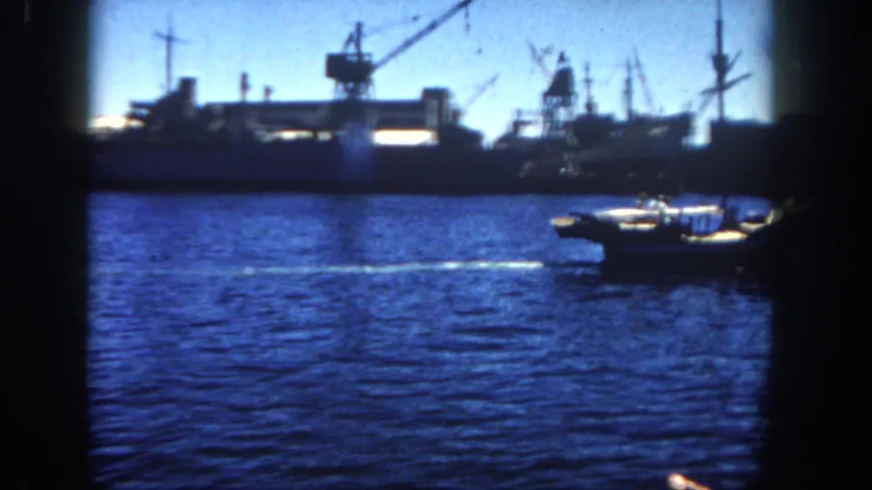 Small motorboat crossing the 1962 San Diego harbor foreground, with docked ships, cranes, and masts along the industrial waterfront behind it.