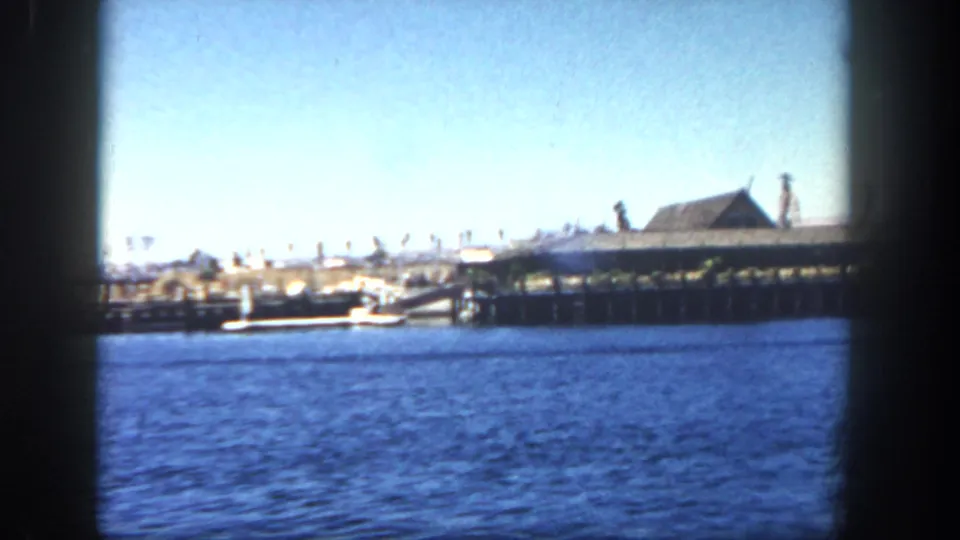 1962 San Diego harbor view from a moving boat, showing blue water, a long pier on pilings, and a low roofed pier building.