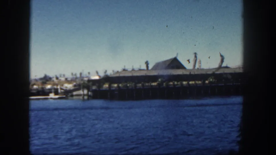 Boat-shot 1962 San Diego waterfront view with a long pier, low building, blue water, and distant palm shapes under a pale sky.