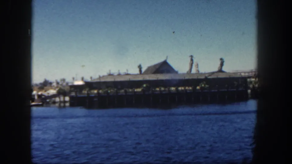 End-frame view of the 1962 San Diego harbor clip, showing rippled water, a pier on dark pilings, and a roofed waterfront structure.