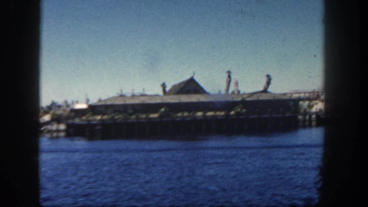 1962 San Diego waterfront view of a large building on a wooden pier across calm blue water, with tall decorative forms on the roofline.