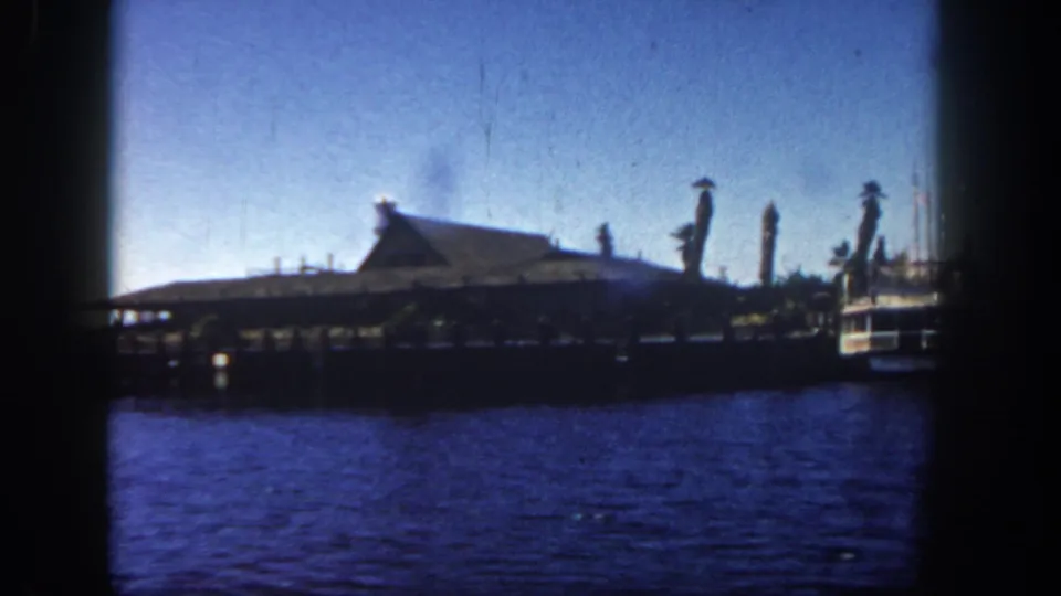 1962 San Diego waterfront frame with a pier on pilings, pointed-roof building, small water tower, and part of a boat at right.