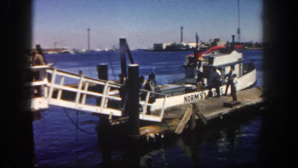 1962 San Diego harbor view of men near a small white boat and wooden dock, seen through soft 8mm frame edges.