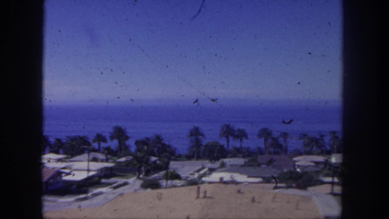 Elevated coastal view with rooftops, palm trees, ocean horizon, blue sky, and dark film edges.