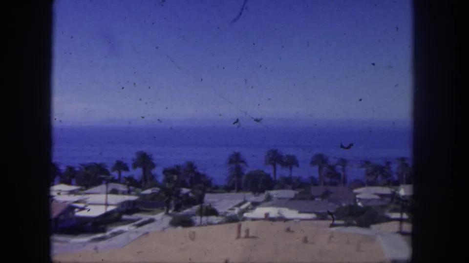 1962 San Pedro home-movie frame showing a distant ocean horizon beyond low houses and palm trees from an elevated viewpoint.