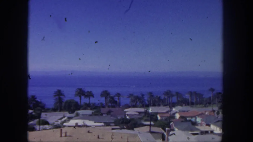 1962 San Pedro coastal view showing low houses, palms, and a blue ocean horizon within visible 8mm frame borders.
