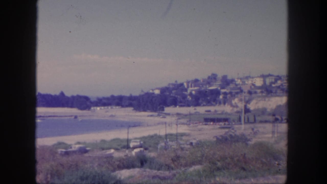 Beach and calm water seen from above, with hillside homes and a dark vertical foreground shape.