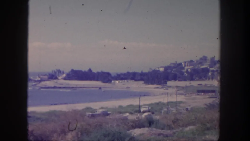 1962 San Pedro frame looking down on shoreline, blue water, hillside houses, low vegetation, cars, and utility poles.