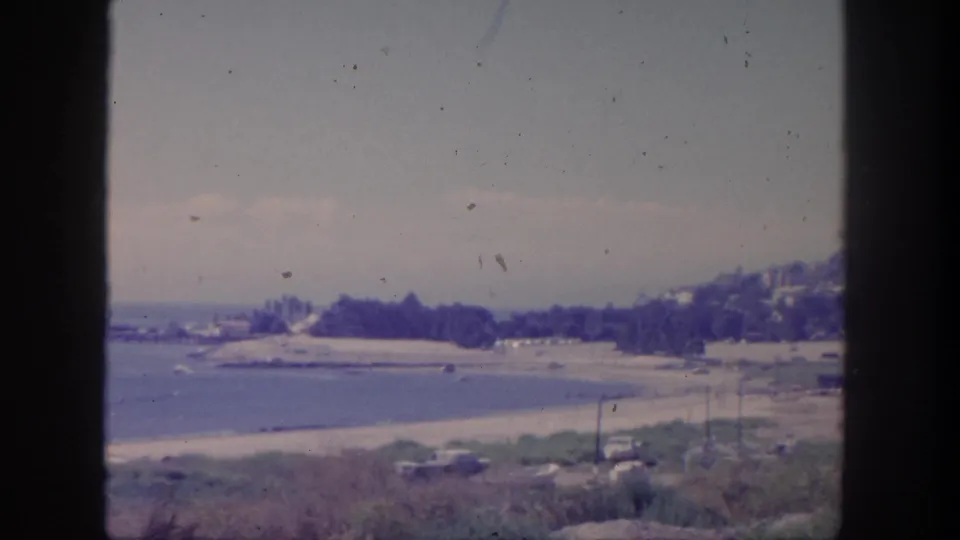 1962 San Pedro home-movie still of a beach and calm water below hillside residences, framed by utility poles and roadside cars.